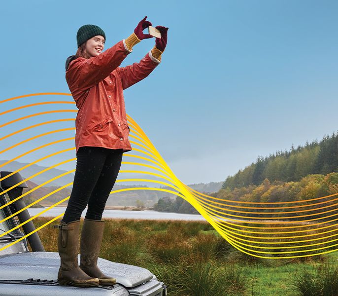 Woman standing on the hood of a car taking a photo of a mountain range with her phone